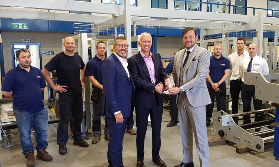 Three males smiling and shaking hands with more employees in the background